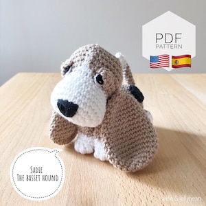 May include: A crocheted stuffed toy basset hound dog named Sadie. The dog is brown and white with black eyes and a black nose. The dog is sitting on a wooden surface. The text "Sadie The Basset Hound" is in a speech bubble.