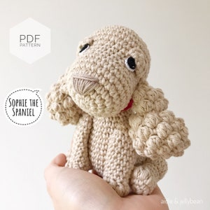 Puede incluir: Un juguete de perro de crochet beige con ojos negros y un collar rojo. El juguete se llama "Sophie the Spaniel" y es un patr&oacute;n PDF.