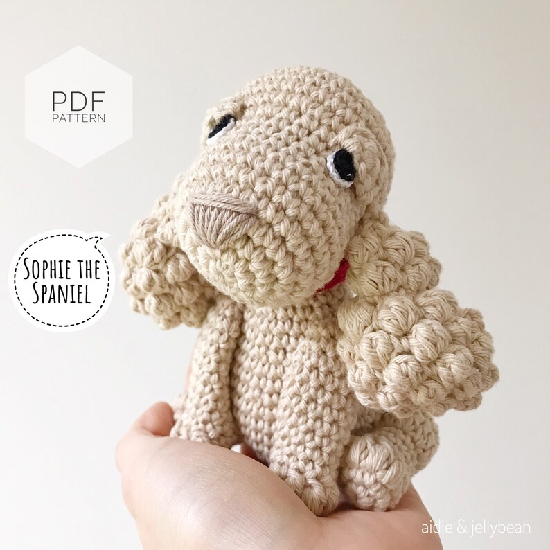 Puede incluir: Un juguete de perro de crochet beige con ojos negros y un collar rojo. El juguete se llama "Sophie the Spaniel" y es un patr&oacute;n PDF.