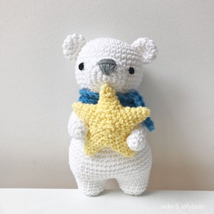 May include: A white crocheted polar bear holding a yellow crocheted star. The bear is wearing a blue scarf.