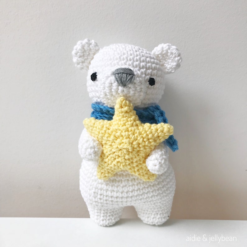 May include: A white crocheted polar bear holding a yellow crocheted star. The bear is wearing a blue scarf.