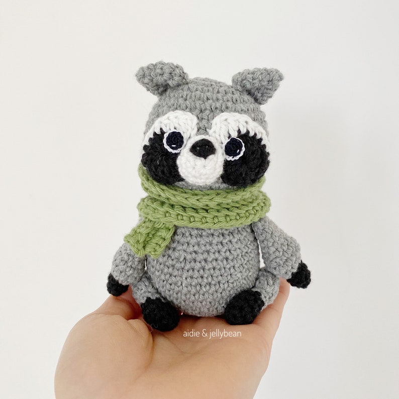 May include: A gray crocheted raccoon wearing a green scarf. The raccoon has black eyes and a black and white face.