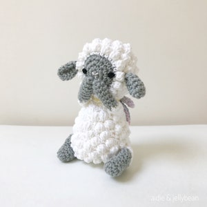 May include: A white crocheted sheep with gray face and legs, sitting with its head down. The sheep has a pink and white ribbon around its neck.