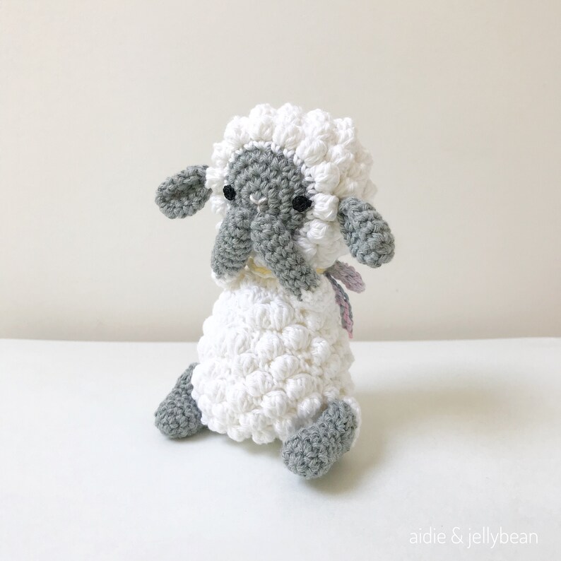 May include: A white crocheted sheep with gray face and legs, sitting with its head down. The sheep has a pink and white ribbon around its neck.