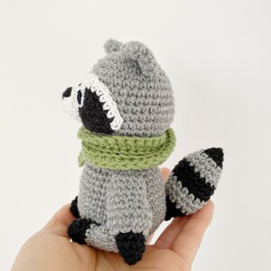 May include: A crocheted raccoon plush toy with a gray body, black and white face, and a green scarf. The toy is sitting in a person's hand.