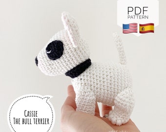May include: White crocheted amigurumi bull terrier dog toy with black eyes and a black collar. The toy is being held in a hand. The text "Cassie The Bull Terrier" is in a speech bubble below the toy. The text "PDF PATTERN" is in a hexagon above the toy. The American and Spanish flags are to the right of the text.
