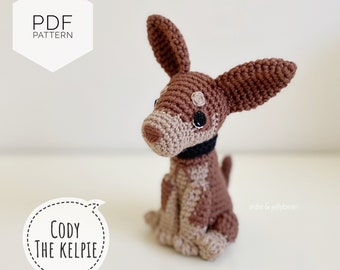 Może przedstawiać: Szydełkowy pies w kolorze brązowym i beżowym z czarnymi oczami i czarnym kołnierzem. Pies siedzi na białym tle. Tekst "Cody The Kelpie" znajduje się w dymku mowy po lewej stronie psa. Tekst "PDF Pattern" znajduje się w sześciokącie w lewym górnym rogu obrazu. Tekst "adie & jellybean" znajduje się w prawym dolnym rogu obrazu.