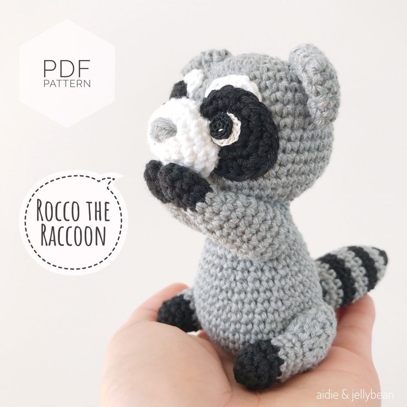 May include: Crochet pattern for Rocco the Raccoon, a gray crocheted stuffed animal with black and white facial features. The pattern is available as a PDF download.