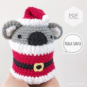 May include: A crocheted Koala Santa figure, featuring a gray koala head with a red Santa hat and a red and white Santa suit. The figure has a black nose and eyes, and a gold-colored belt buckle. The text "Koala Santa" is in a speech bubble.