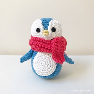 May include: A crocheted penguin wearing a red scarf. The penguin is blue and white with black eyes and a yellow beak.