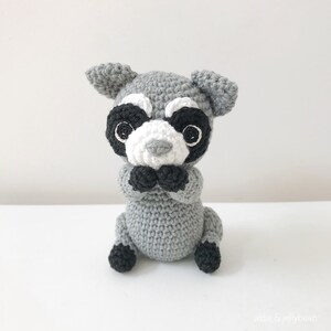 May include: A crocheted gray raccoon plush toy with black eyes, a white nose, and black ears. The toy is sitting with its paws in front of it.