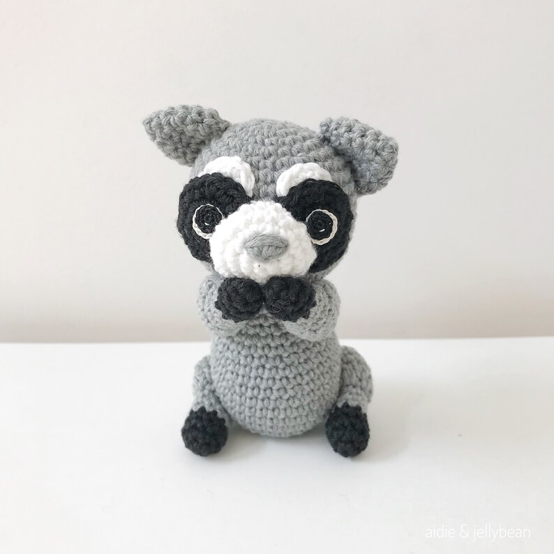 May include: A crocheted gray raccoon plush toy with black eyes, a white nose, and black ears. The toy is sitting with its paws in front of it.