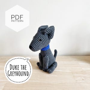 May include: A crocheted greyhound dog with a blue collar. The dog is sitting, with its ears perked up. The image includes text that says "Duke the Greyhound" and "PDF Pattern". The dog is handmade and sits on a wooden surface.