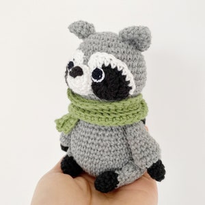 May include: A crocheted raccoon plush toy wearing a green scarf. The toy has black eyes, a black nose, and black and white markings on its face.