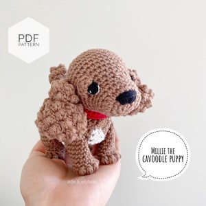 May include: A crocheted brown puppy with a red collar and white patch on its chest. The puppy has black eyes and a black nose. Text on the image reads "PDF PATTERN" and "MILLIE THE CAVOODLE PUPPY".