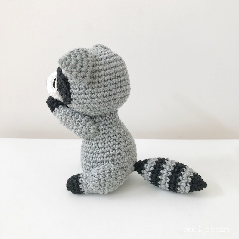 May include: A gray crocheted raccoon amigurumi toy with black stripes on its tail. The toy is sitting on a white surface.