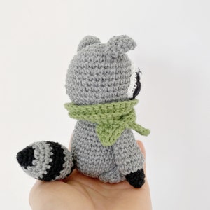 May include: A gray crocheted raccoon with a green scarf and a black and white striped tail.