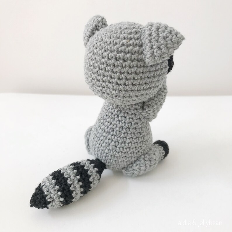 May include: A crocheted raccoon amigurumi toy with a gray body and a black and gray striped tail. The toy is sitting on a white surface.