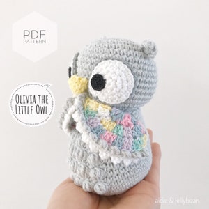 May include: A crocheted gray owl with a colorful scarf. The owl has large white eyes with black pupils and a yellow beak. The scarf is made of a variety of colors, including pink, blue, yellow, and green. The owl is sitting on a hand.