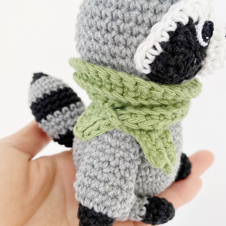 May include: A crocheted raccoon toy wearing a green scarf and a white crocheted hat. The toy is gray with black stripes on its tail.