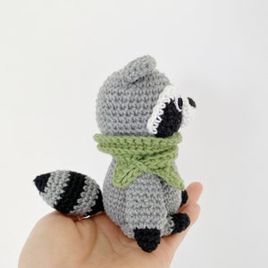 May include: A crocheted raccoon plush toy wearing a green scarf. The toy has black eyes, a white nose, and a black and white striped tail.