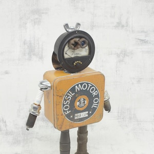 Puede incluir: Un robot hecho de materiales reciclados, incluyendo una lata de hojalata con las palabras "Fossil Motor Oil" impresas en ella, un medidor vintage y bujías. El robot tiene una cabeza redonda con dos ojos azules y una perilla plateada en la parte superior.