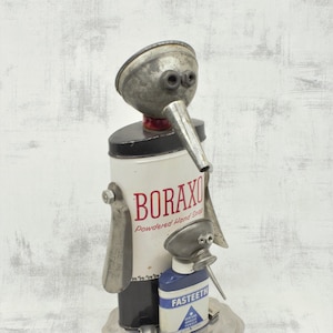 May include: A whimsical sculpture made from recycled materials, featuring a figure with a funnel head and a Boraxo powdered hand soap can body. The figure is holding a bottle of  Fastteeth denture powder. The sculpture is standing on a silver tray.