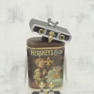 May include: A robot made from recycled materials, including a Hershey's Cocoa tin, with a silver camera lens on top and two forks for legs.