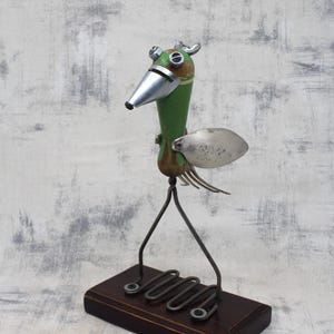 Puede incluir: Una escultura de pájaro caprichosa de color verde y plateado hecha con materiales reciclados. El pájaro tiene un pico largo, ojos grandes y un ala hecha de una cuchara. Se encuentra sobre una base de madera con patas de metal.