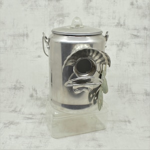 May include: A silver metal birdhouse made from a repurposed coffee pot with a clear plastic base. The birdhouse has a round opening and a decorative butterfly design.
