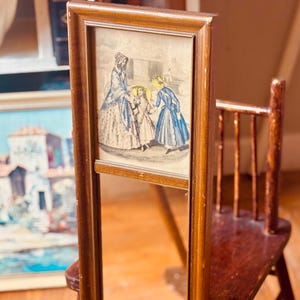 May include: A vintage, rectangular mirror with a wooden frame. The top section features a framed print of figures in period clothing. The mirror is propped up on a wooden floor next to a small wooden chair.