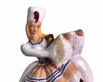 Puede incluir: Una figurita de cerámica de una mujer con un vestido blanco y una falda con estampado rojo, azul y amarillo. Lleva un sombrero blanco con una pluma roja y sostiene un abanico.