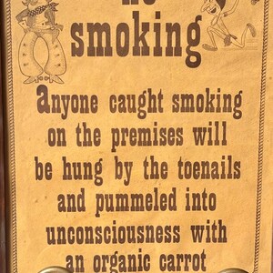 May include: A vintage-style sign with the words "no smoking" in large, bold letters. The sign features cartoon illustrations of a cowboy and a Native American. The text below warns of consequences for smoking on the premises.