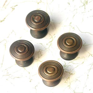 May include: Four antique bronze cabinet knobs with a circular, ridged design. The knobs have a warm, aged patina, suggesting they are vintage or vintage-inspired. The knobs are likely used for drawers or cabinet doors.