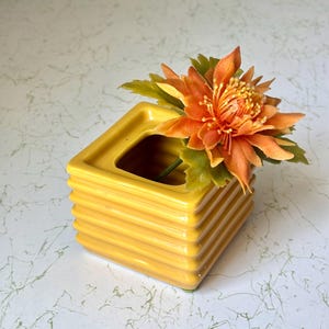 May include: A square, mustard yellow ceramic vase with a ribbed, tiered exterior design. The vase features a wide, open top and contains a single artificial orange flower with green leaves extending from the center of the opening.