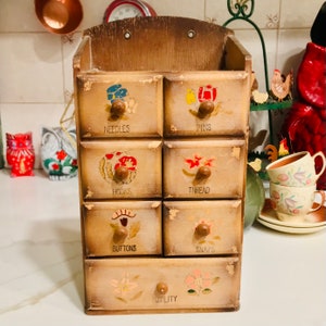 Puede incluir: Una caja de madera con siete cajones, cada uno con una etiqueta diferente y un diseño pintado. Las etiquetas son "Agujas", "Alfileres", "Ganchos", "Hilo", "Botones", "Broches" y "Utilidad".