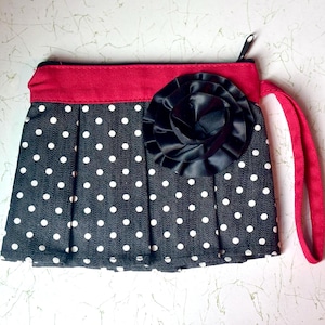 Puede incluir: Un clutch de lunares blancos y negros con la parte superior y el asa rojas. El clutch presenta una gran flor negra y un cierre de cremallera. El diseño plisado añade un toque de elegancia a este accesorio.