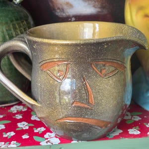 Könnte beinhalten: Ein handgefertigter Keramikkrug mit einem Gesichtsdesign. Der Krug ist hauptsächlich olivgrün mit einem beigefarbenen Innenraum und braunen Akzenten, die die Gesichtszüge bilden. Er hat einen gebogenen Griff und einen Ausguss.