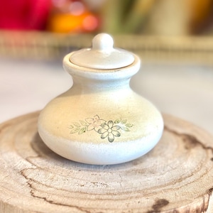 May include: A small, off-white ceramic jar with a lid, decorated with a floral design. The jar is sitting on a wooden surface. The lid has a small knob on top. The floral design features green leaves and a pink flower.