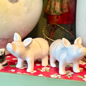 Könnte beinhalten: Zwei kleine, glänzende, weiße Keramik-Schweinefiguren. Die Schweine stehen, mit aufgestellten Ohren und nach vorne gerichteten Schnauzen. Sie werden auf einer rot geblümten Oberfläche präsentiert. Links ist eine kleine schwarz-weiße Katzenfigur zu sehen.