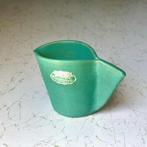 May include: A teal-colored ceramic ashtray with a unique, curved design. The ashtray features a small, oval sticker with the text "Rosemeade" in a script font. The ashtray's shape is reminiscent of a small pitcher or scoop.