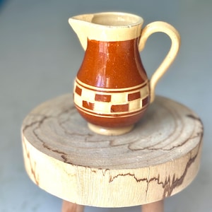 May include: A small, ceramic pitcher with a brown body and a cream-colored rim and handle. The pitcher features a decorative band of cream and brown checkered squares around its midsection. It is displayed on a small, round wooden stool.