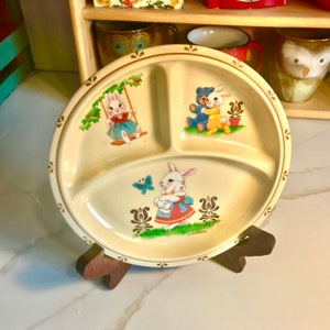 May include: A divided ceramic plate with a cream background and brown trim. The plate features three sections with illustrations of bunnies and a teddy bear. The plate is on a wooden stand.
