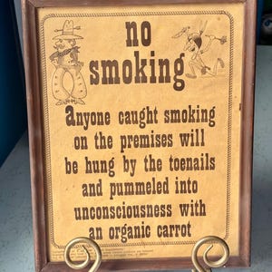 May include: A framed "No Smoking" sign with humorous text and cartoon illustrations. The sign has a brown wooden frame and is propped up by a gold-colored metal stand. The text warns of harsh consequences for smoking.