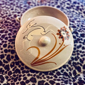 May include: A ceramic serving dish with a lid. The lid features a hand-painted design of a deer with a flower and leaves in shades of brown, orange, and black on a light gray background.