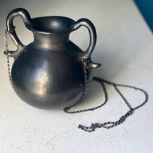 May include: A dark grey ceramic vessel with two arched handles and a small spout on each side. A delicate chain is attached to each spout, and hangs down. The vessel has a smooth, reflective surface and a wide opening.