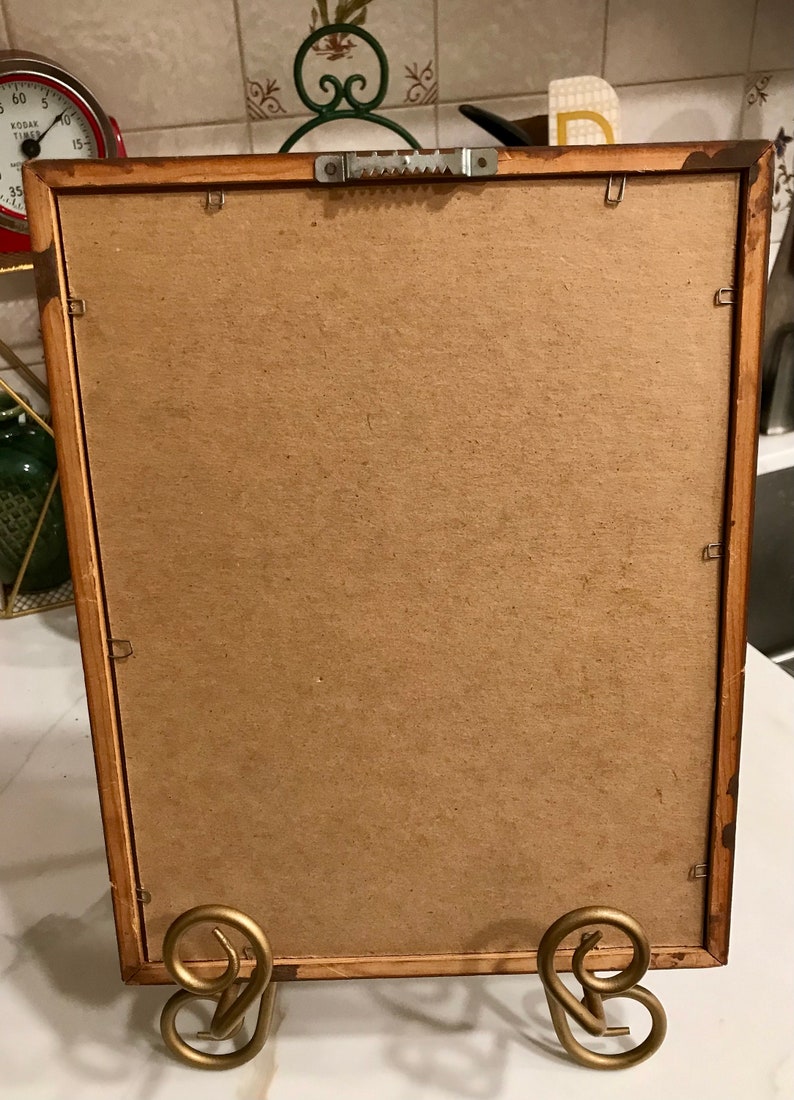 May include: A rectangular wooden picture frame with a cardboard backing. The frame is supported by a gold-colored metal stand. The back of the frame has a sawtooth hanger for wall mounting.