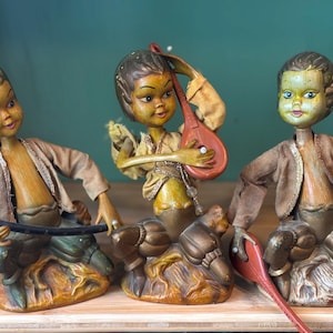May include: Three vintage wooden figurines of children playing music. The figurines are painted in shades of brown, yellow, and gold. One child plays a lute, another plays a flute, and the third plays a tambourine.