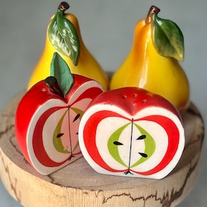 May include: A set of ceramic fruit-shaped salt and pepper shakers. Two yellow pear-shaped shakers with green leaves are behind two red apple-shaped shakers. The apple shakers have a white and green interior design.