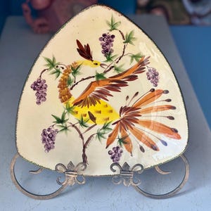 May include: A vintage, triangular-shaped decorative plate with a hand-painted bird design. The bird is yellow, brown, and orange, perched among green leaves and purple grapes. The plate has a cream-colored background and is displayed on a decorative metal stand.